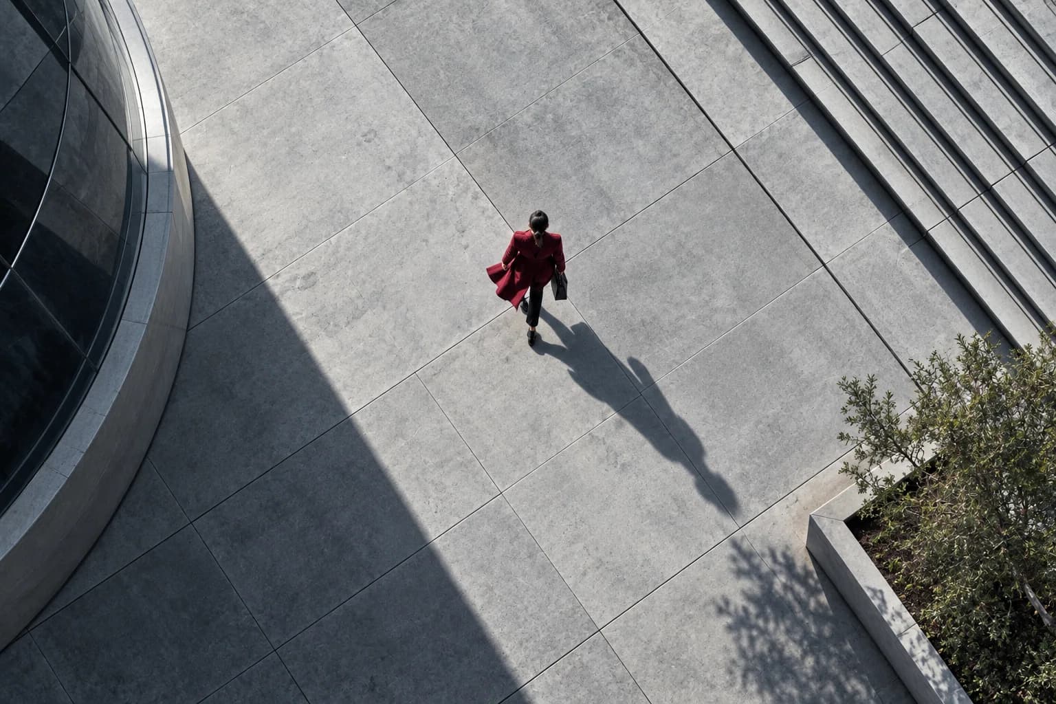 Overhead shot of a burgundy-coated figure and shadow on a pale concrete plaza